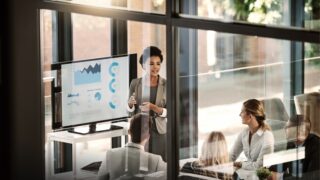 woman of color presenting to room of people in windowed office